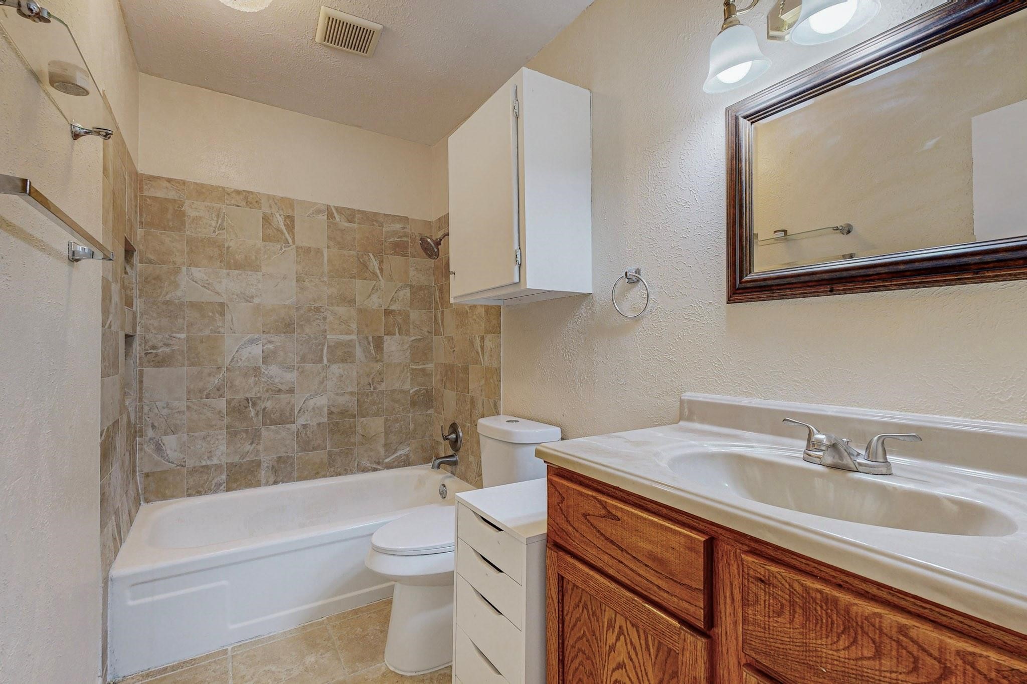 Bathroom with tan tiled tub surround, toilet, and wood vanity with white sink.