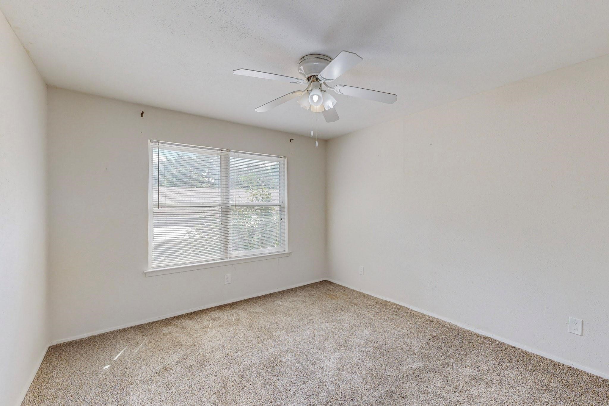 Empty carpeted bedroom with a ceiling fan and a wide window fitted with blinds.