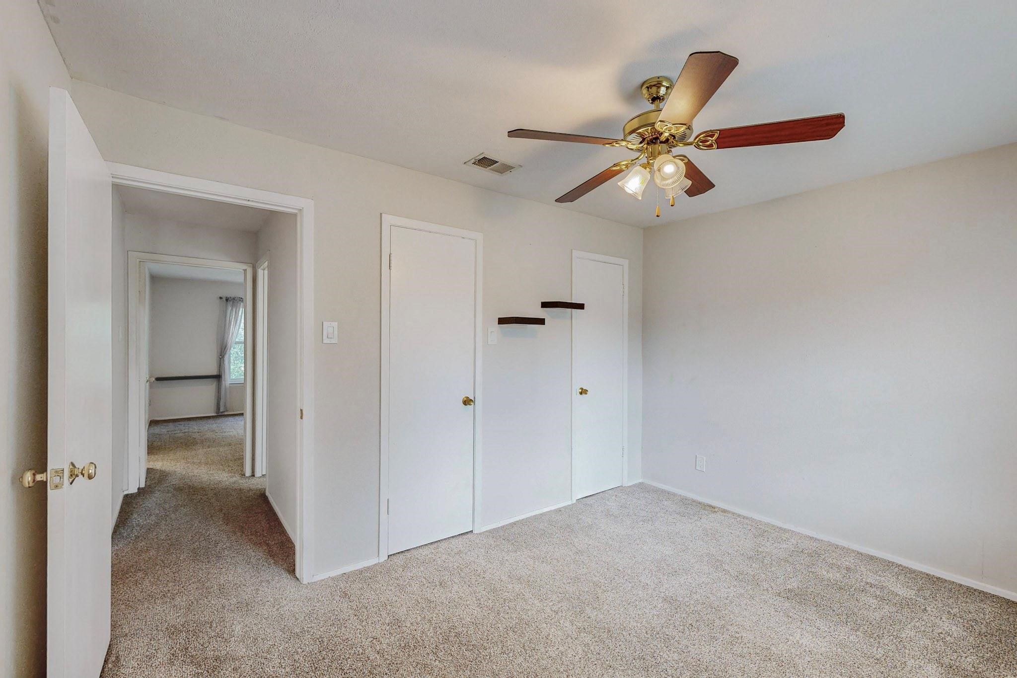 Empty carpeted bedroom with a ceiling fan, two closet doors, and floating shelves.