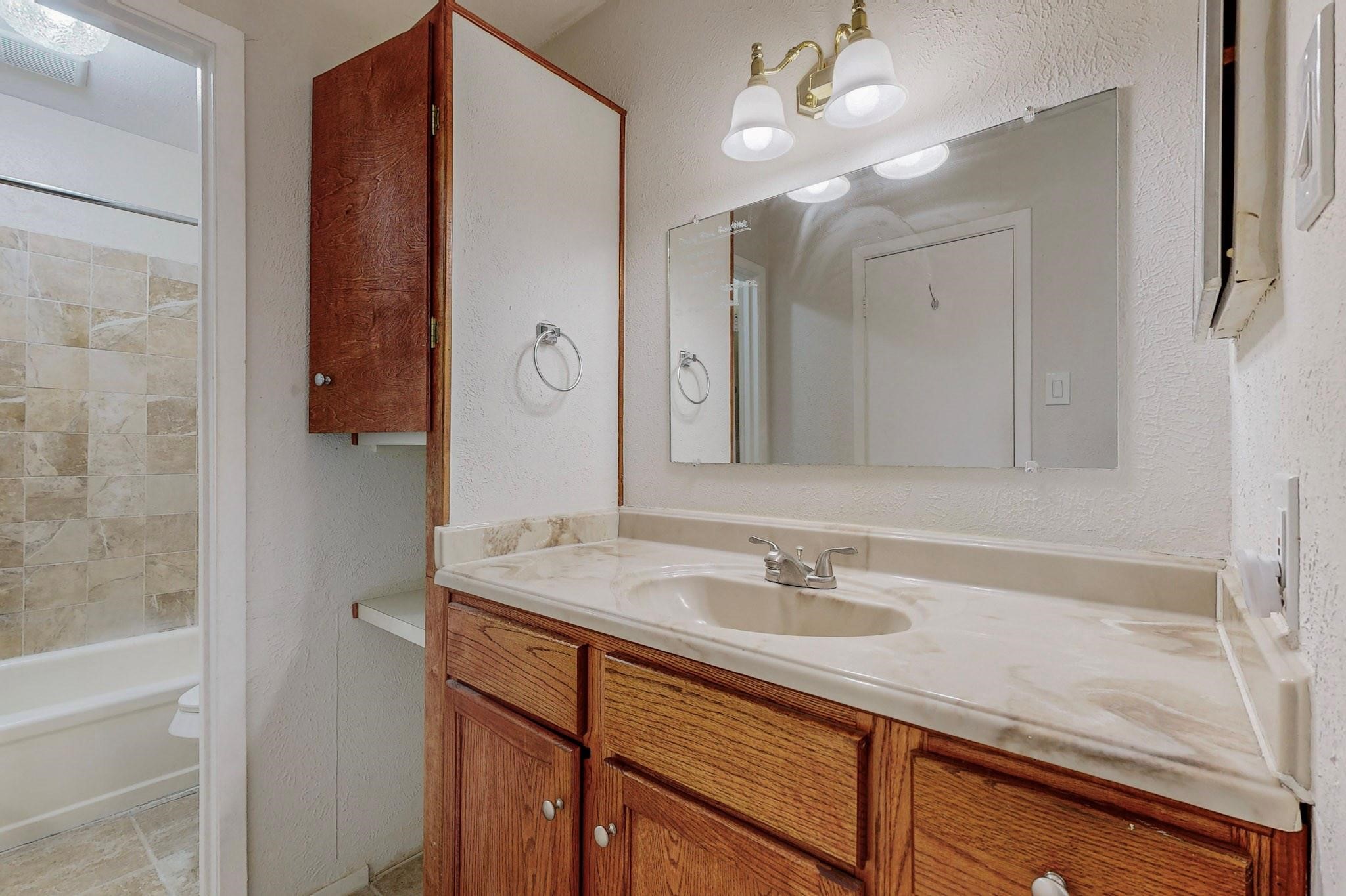 Bathroom vanity with wood cabinets and a tiled tub-shower beside it.