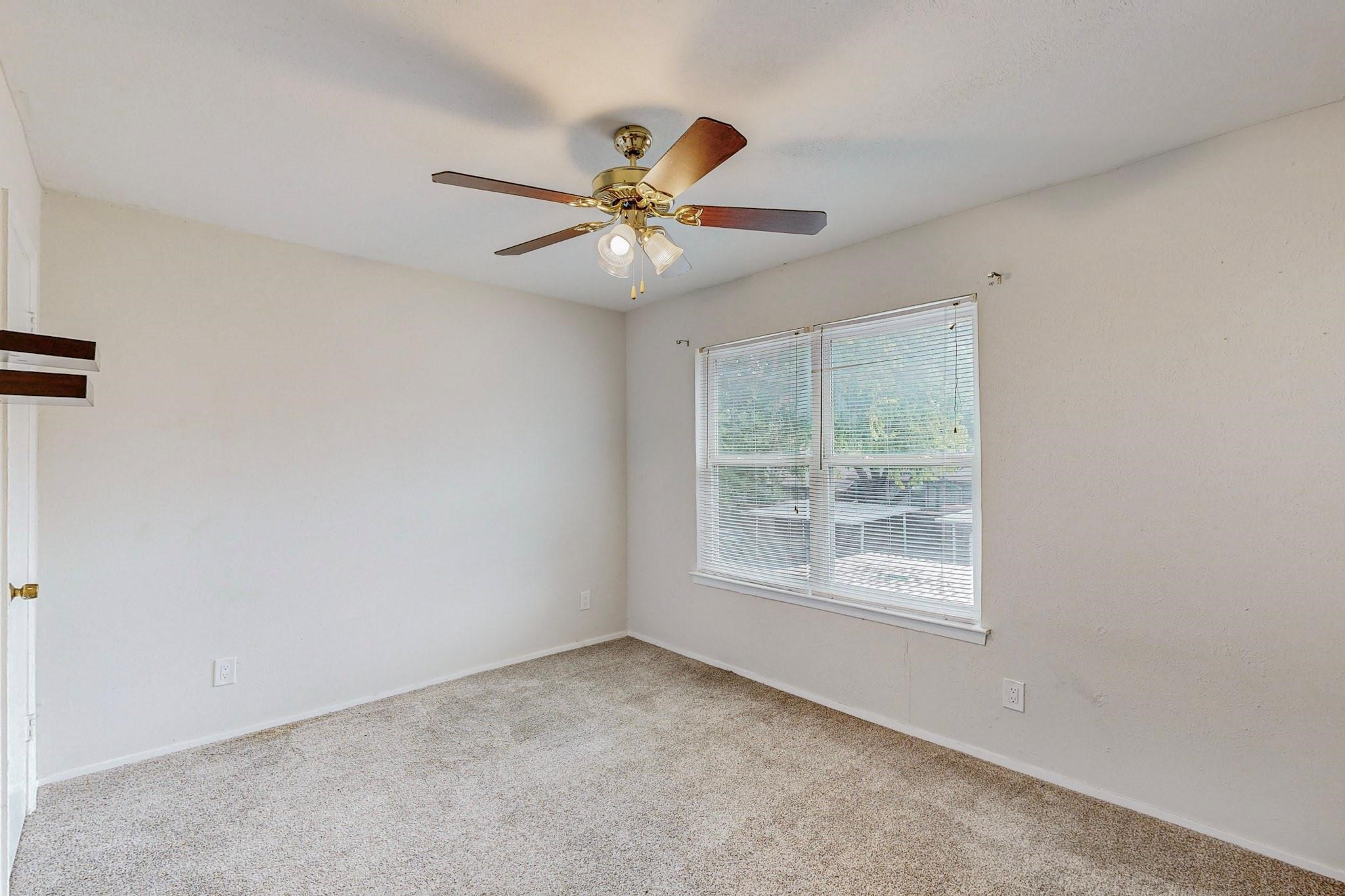 Empty bedroom with beige carpet, a brass ceiling fan, and a wide window with white blinds.