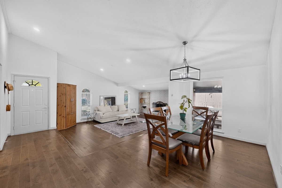 Open-plan living and dining room with vaulted ceiling, dark wood floors, and white brick fireplace.