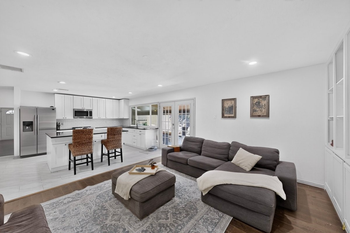 Open-plan living room with a gray sectional and a white kitchen with woven bar stools.
