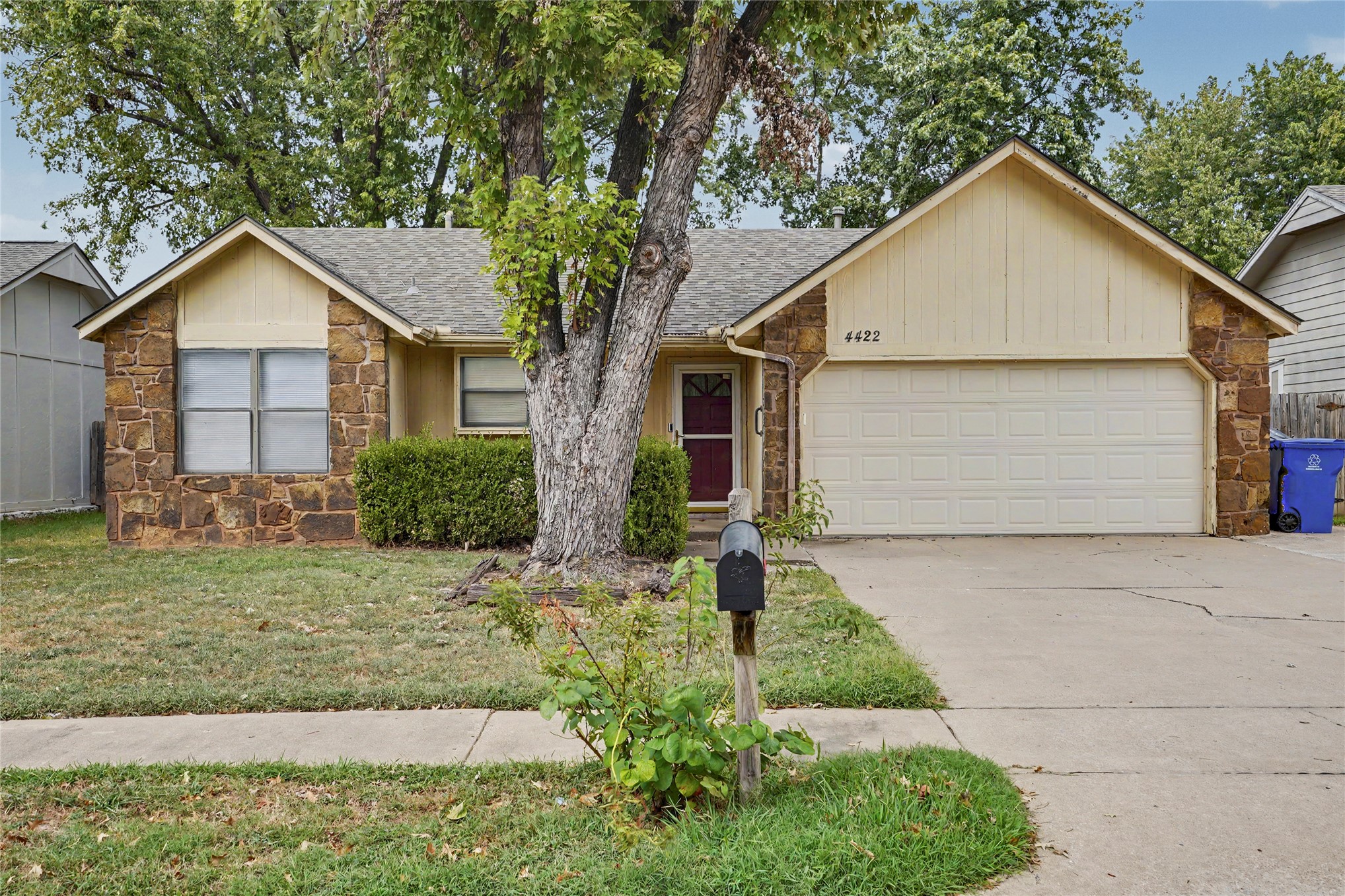 Single-story tan house with a stone facade, attached garage, and large tree in the front yard.