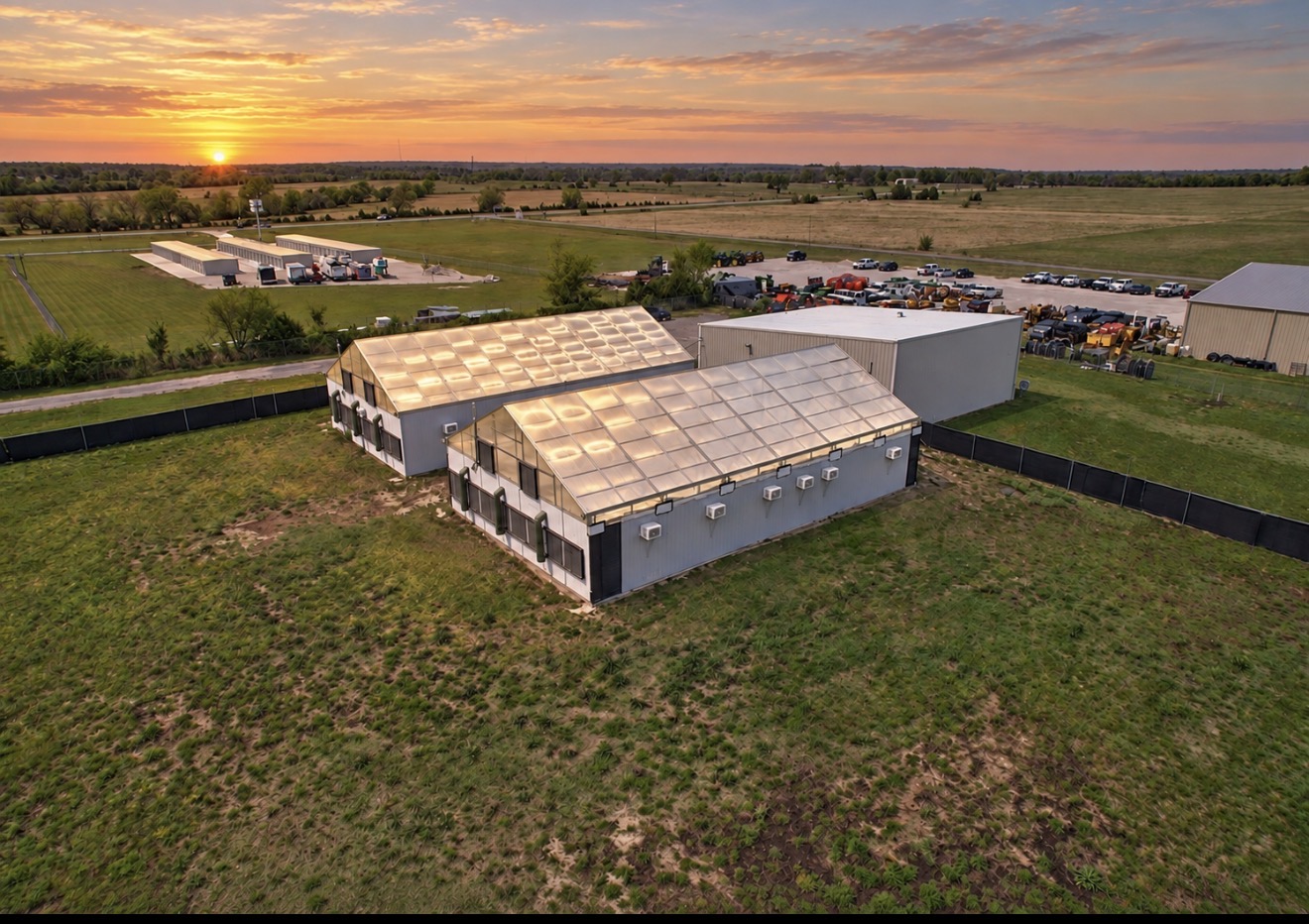 Aerial view of greenhouse buildings and open fields beneath a vivid sunset sky.