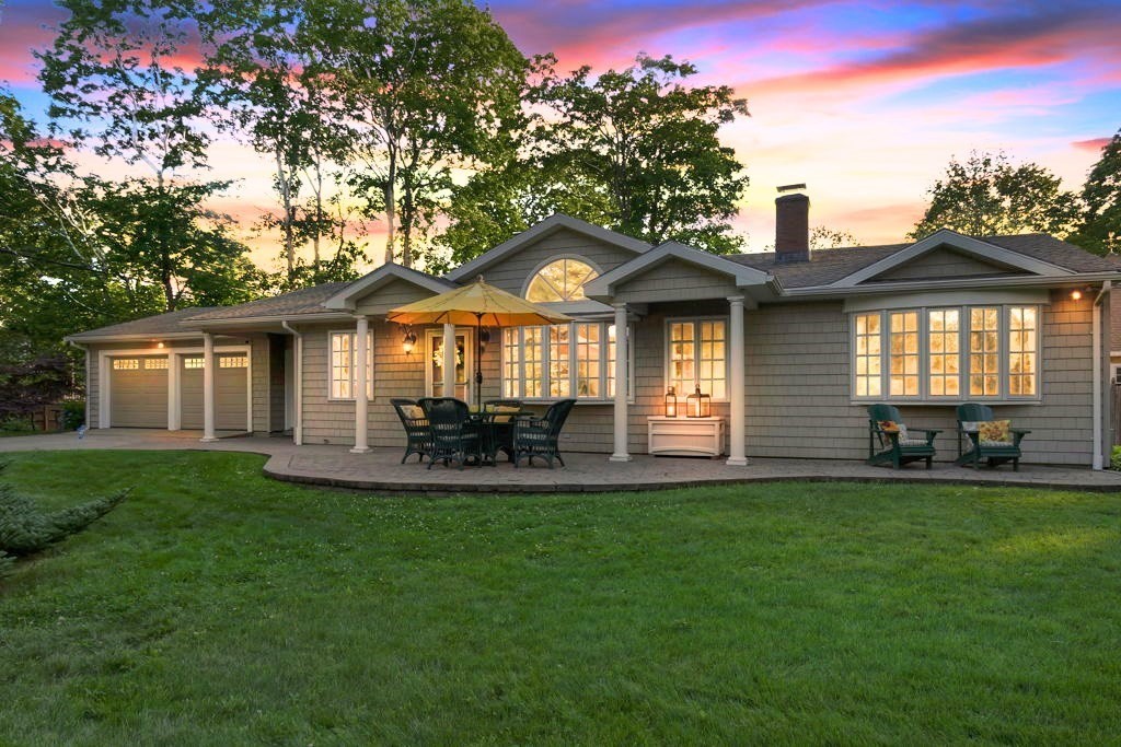 Gray shingle-sided home with an illuminated patio and broad lawn beneath a colorful dusk sky.