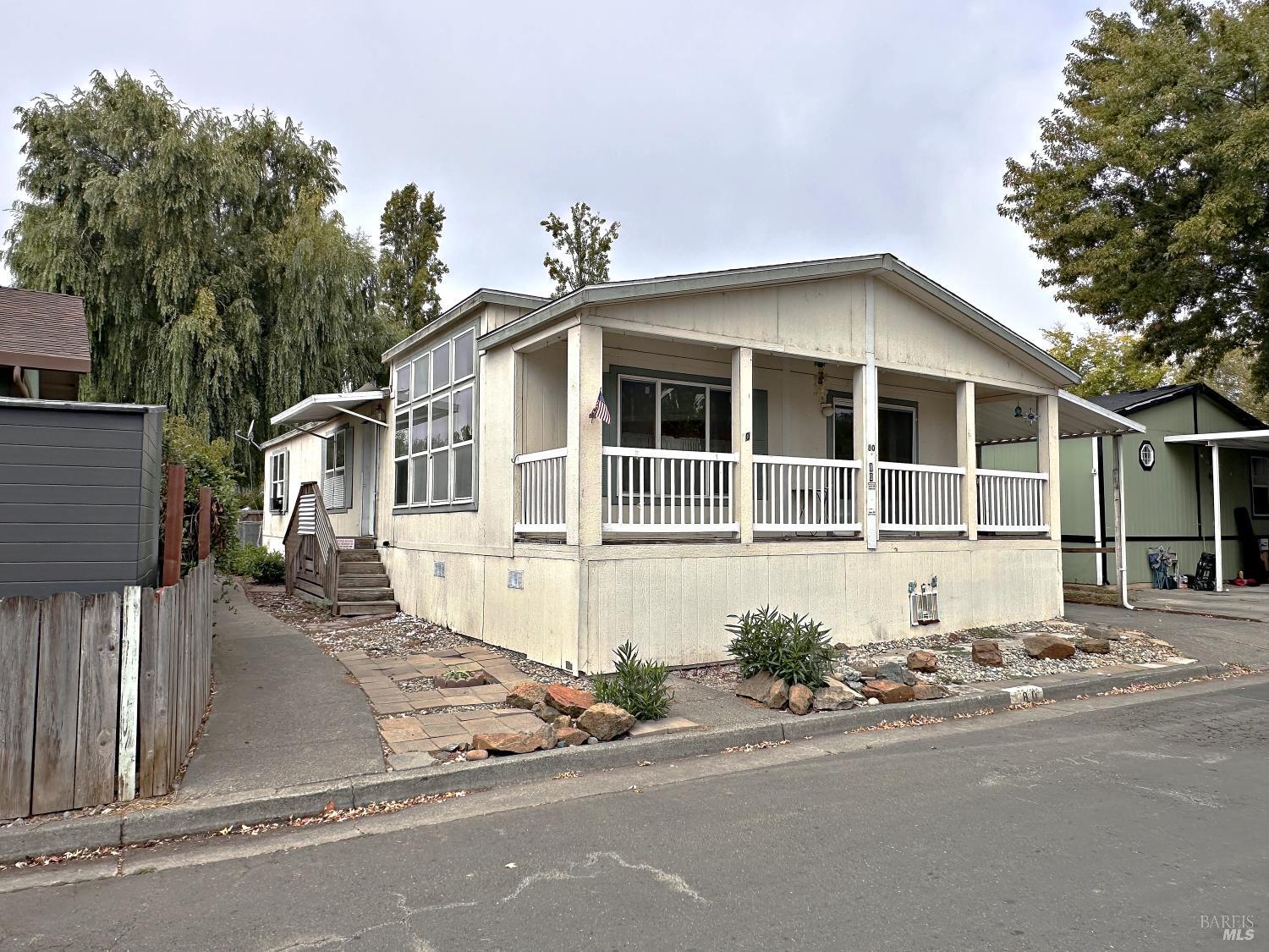 Light-colored manufactured home with a covered porch and white railings beside a small rock garden.