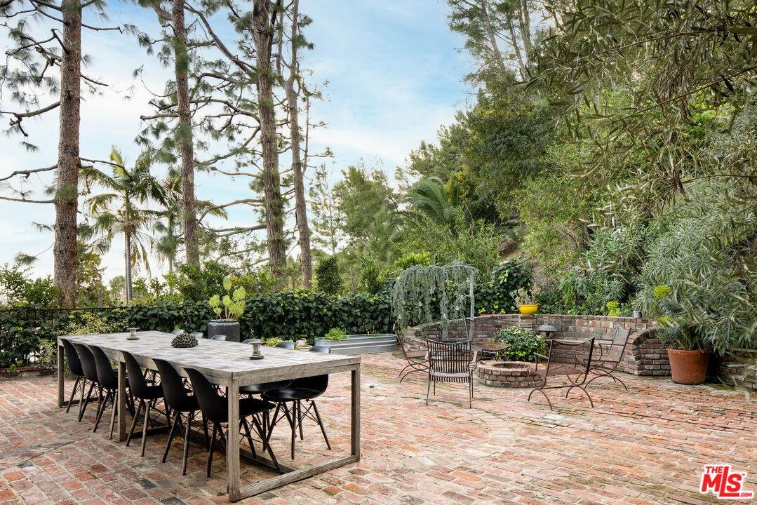 Brick patio with a long outdoor dining table, metal seating, raised planters, and mature trees.