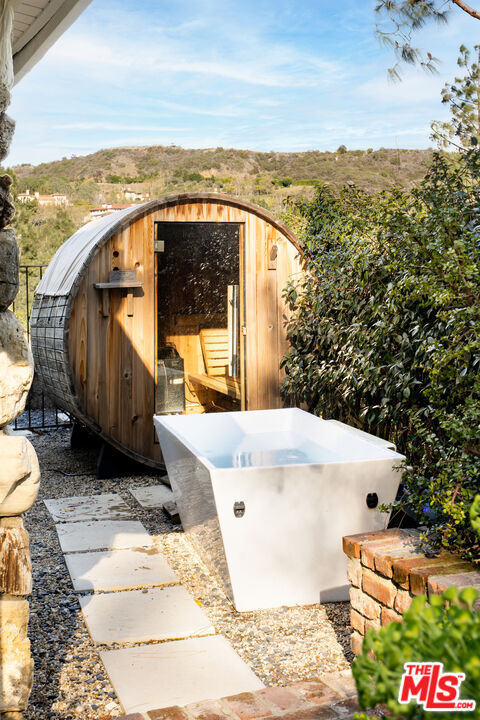 Outdoor soaking tub beside a barrel-shaped wooden sauna, with rolling hills in the distance.