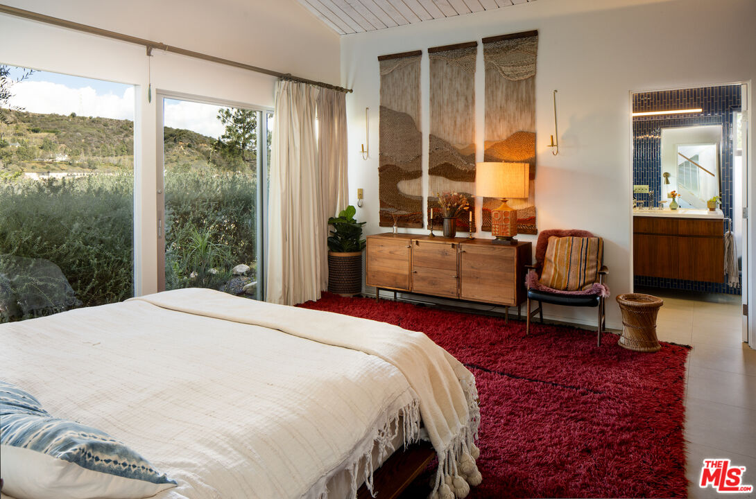 Bedroom with a large bed, red rug, woven wall art, and sliding doors overlooking a green hillside.