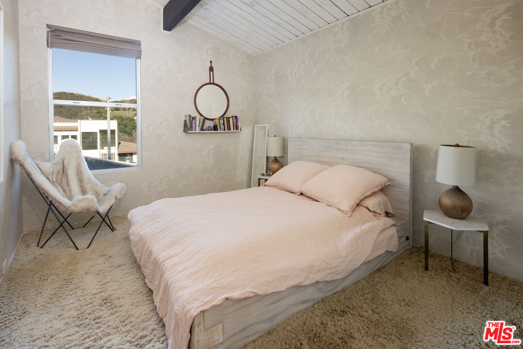 Bedroom with pale pink bedding, a white plank ceiling, and a window overlooking hills.