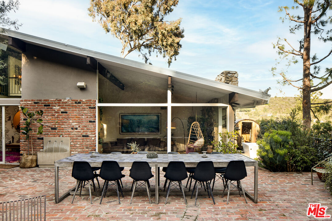Modern home with glass walls and a brick patio dining area beneath mature trees.