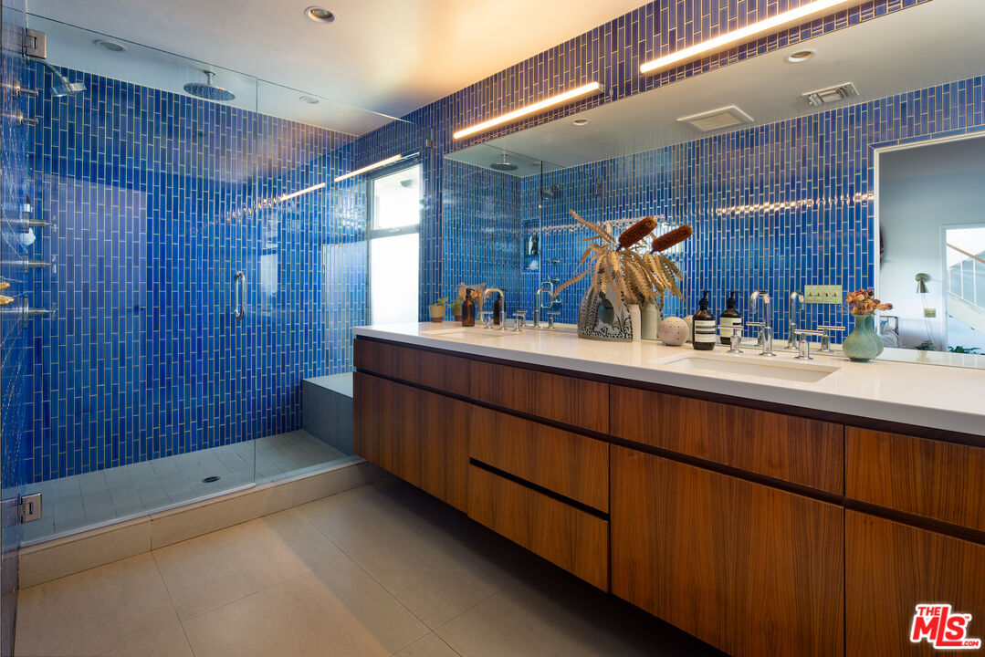 Bathroom with a blue-tiled shower and long walnut vanity beneath a large mirror.