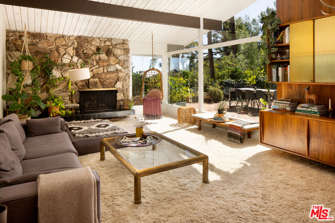 Living room with a stone fireplace, broad garden-facing windows, and mid-century wood cabinetry.
