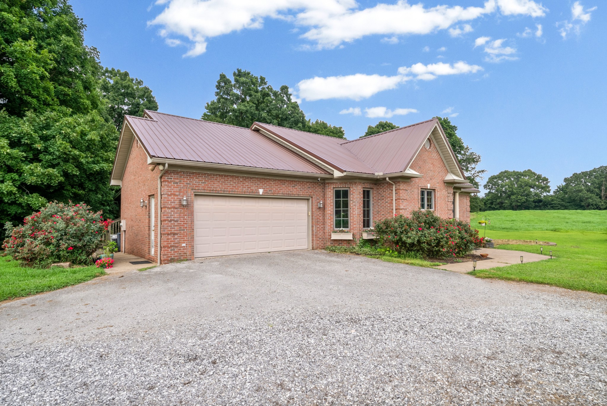 Red brick house with a metal roof, two-car garage, gravel driveway, and flowering shrubs.