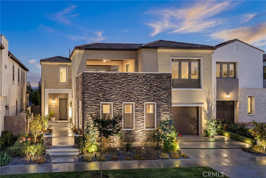 Two-story contemporary home with a stone facade, landscaped entry, and illuminated windows at dusk.