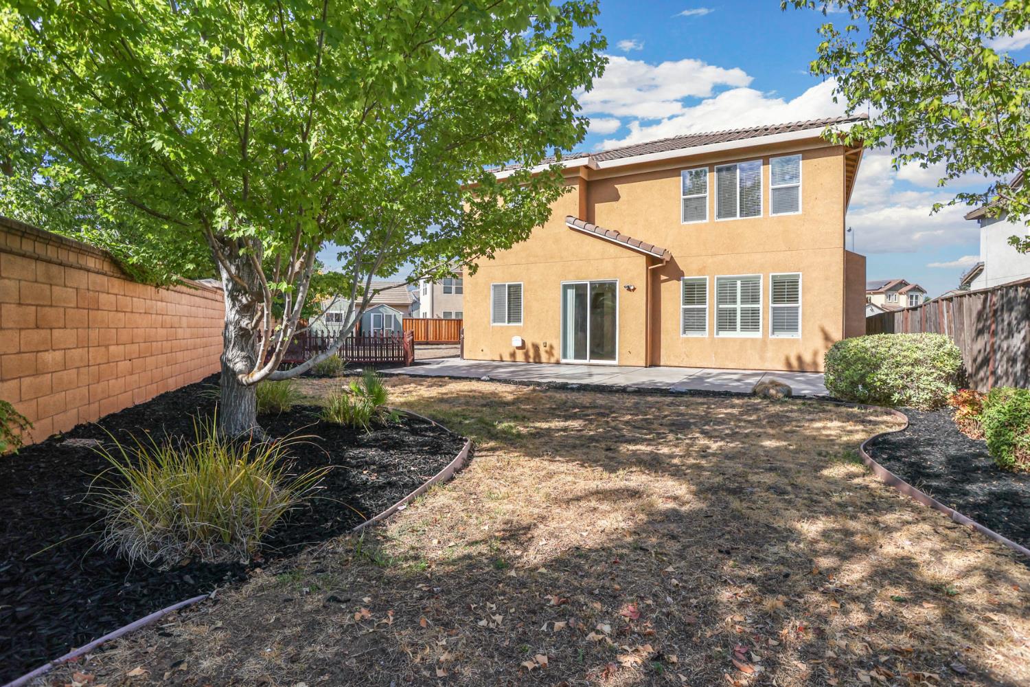 Rear of a two-story tan stucco home with a patio, mature trees, and a fenced backyard.
