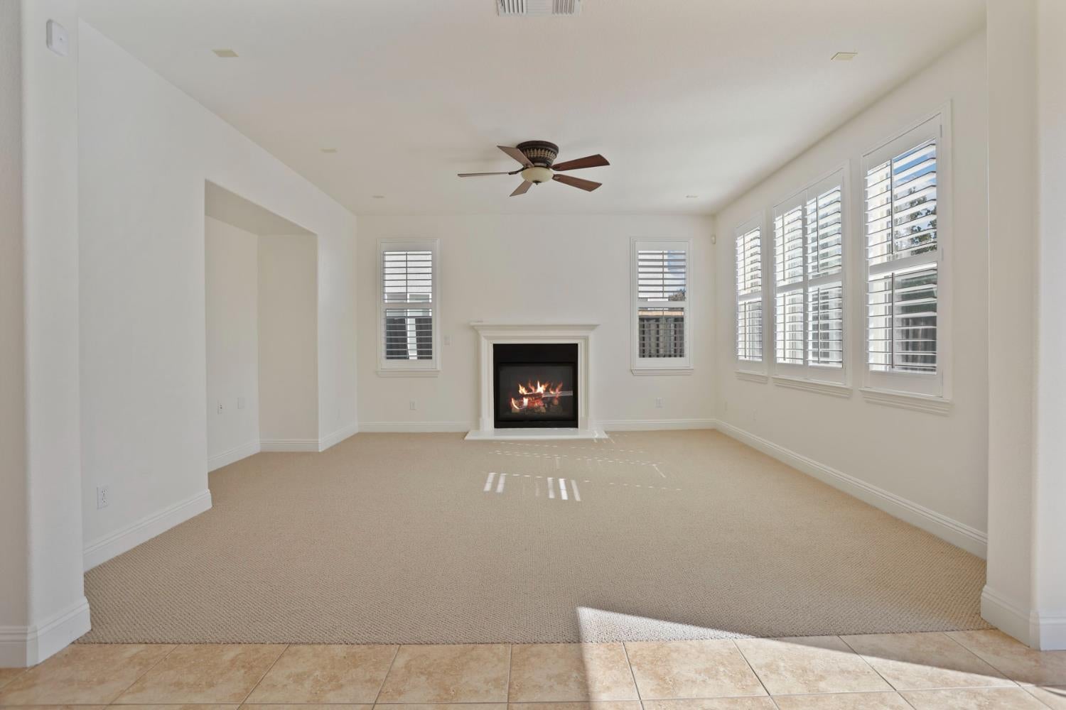 Vacant living room with a fireplace, ceiling fan, and shuttered windows.