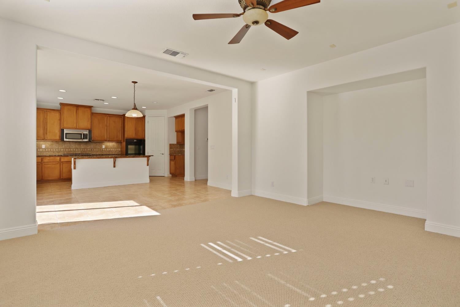 Vacant living room with beige carpet, ceiling fan, and an adjoining wood-cabinet kitchen.