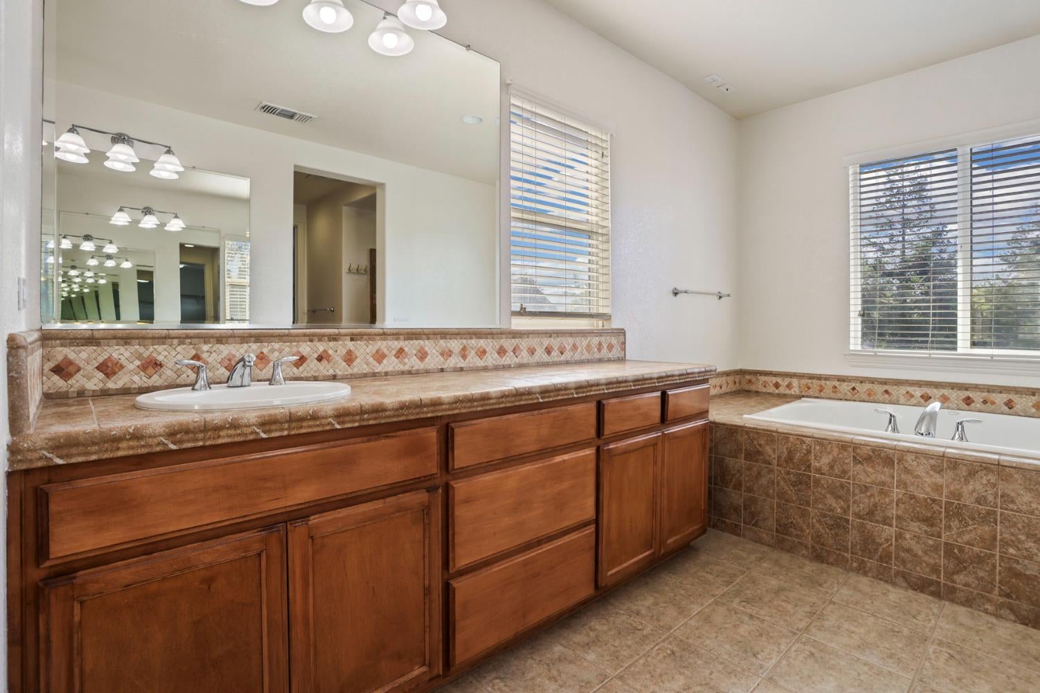 Bathroom with a wood vanity and large mirror beside a tiled bathtub and bright windows.
