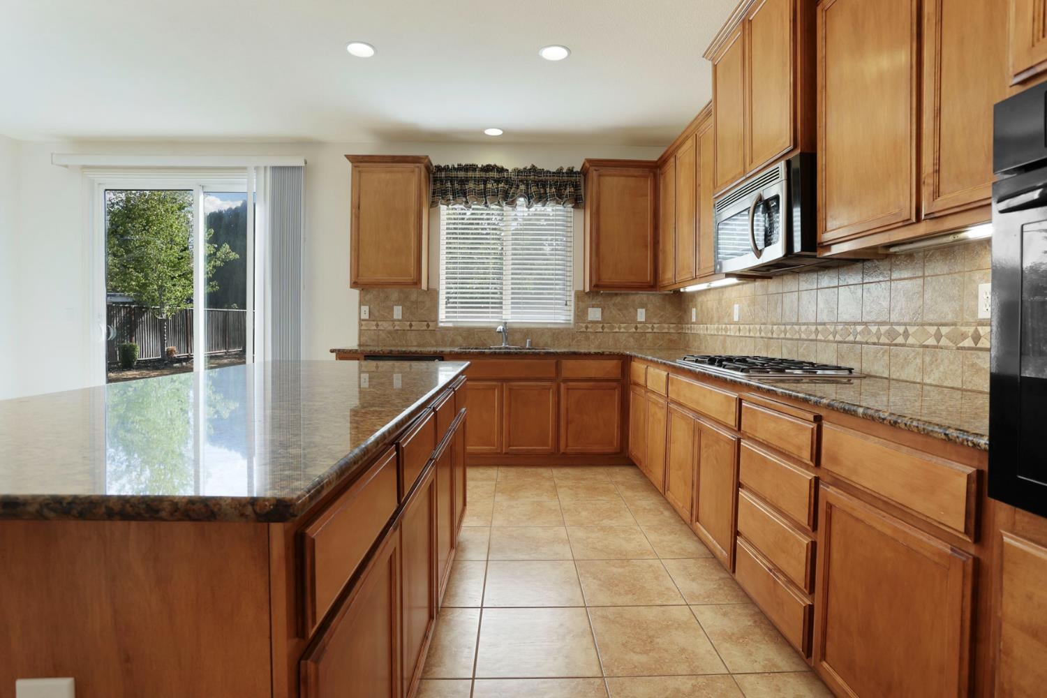 Kitchen with warm wood cabinetry, granite island, tan tile, and a gas cooktop.
