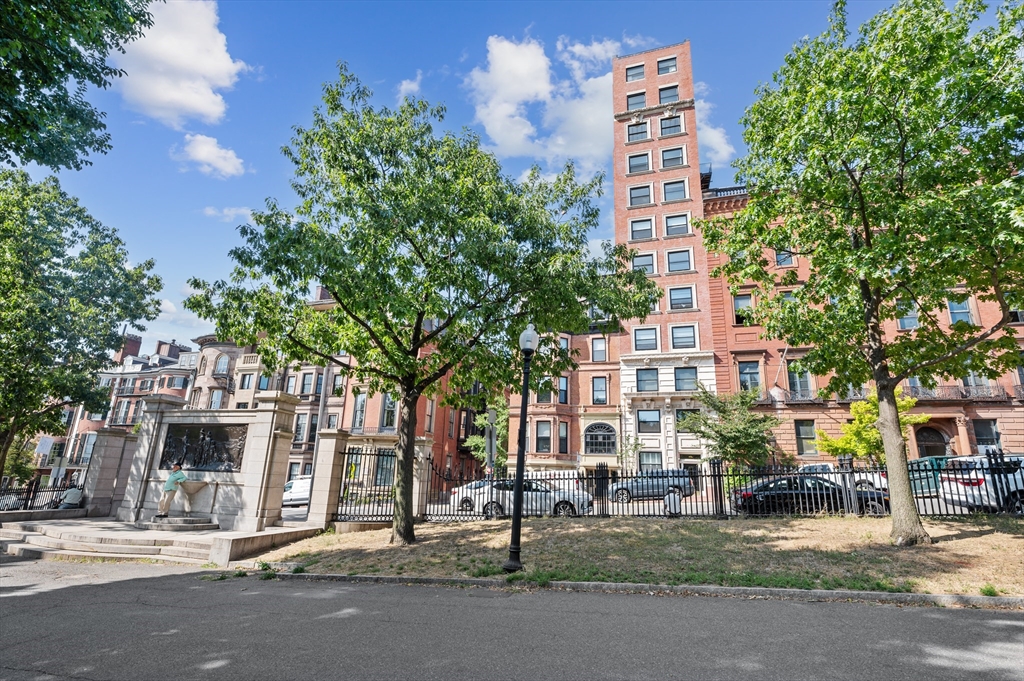 Red-brick apartment building behind an iron fence, framed by leafy trees and neighboring historic buildings.