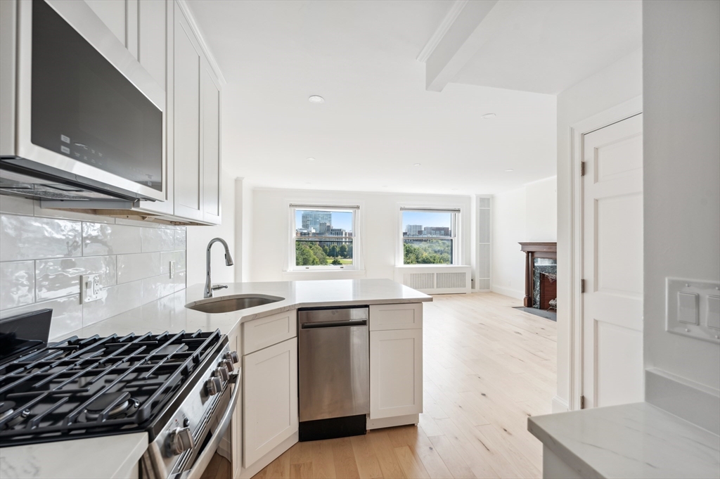 Open-plan kitchen with stainless steel appliances, pale wood floors, and windows overlooking greenery and city buildings.
