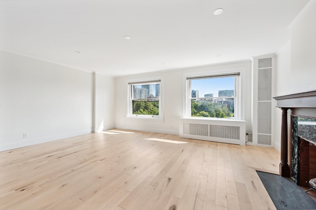 Vacant living room with pale hardwood floors, two city-view windows, and a dark marble fireplace.