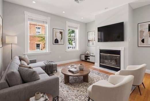 Staged living room with a gray sectional, white armchairs, and a fireplace beneath a wall-mounted TV.
