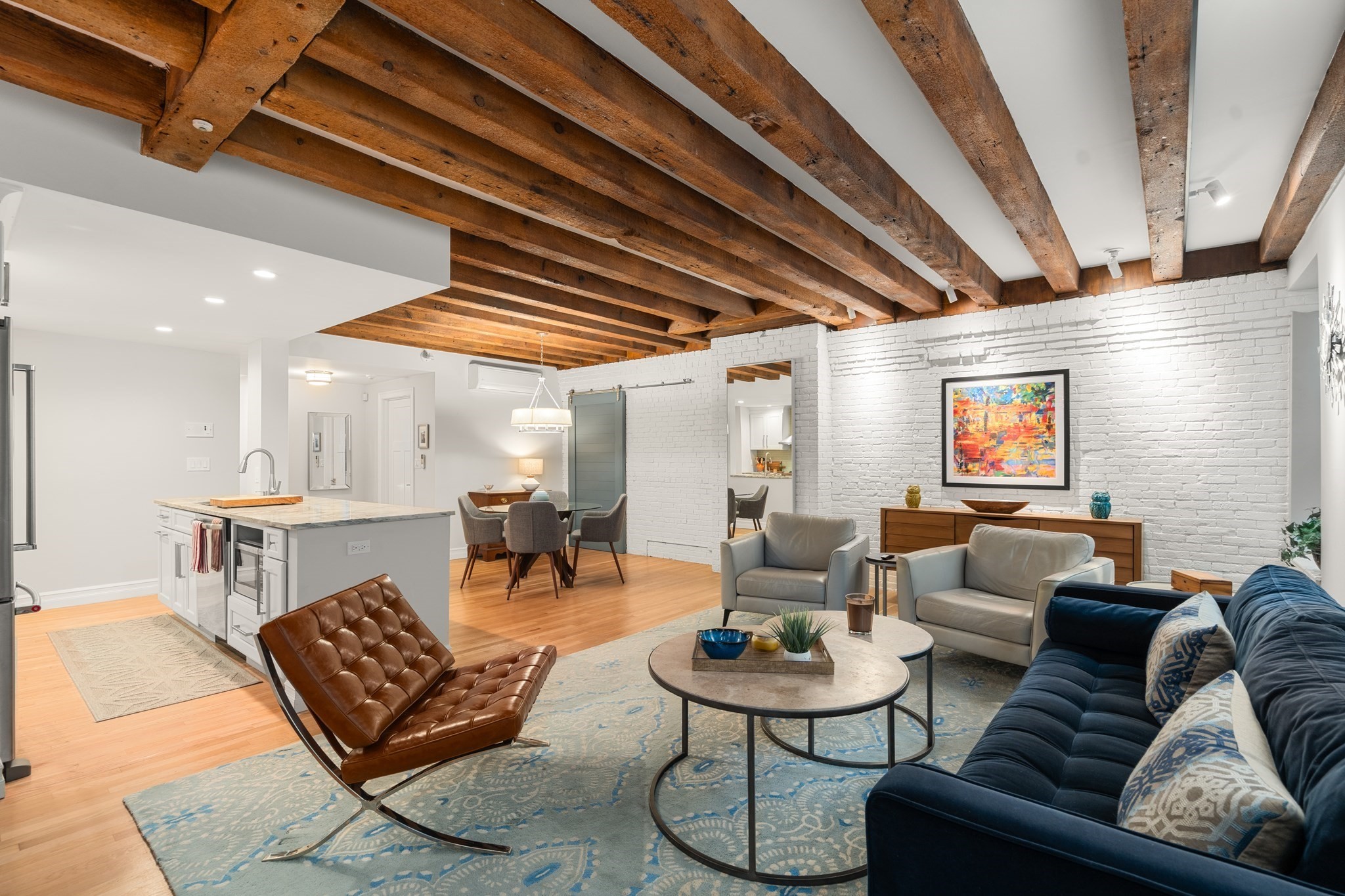 Open-plan loft living room with exposed wooden beams, white brick walls, and an adjoining kitchen and dining area.
