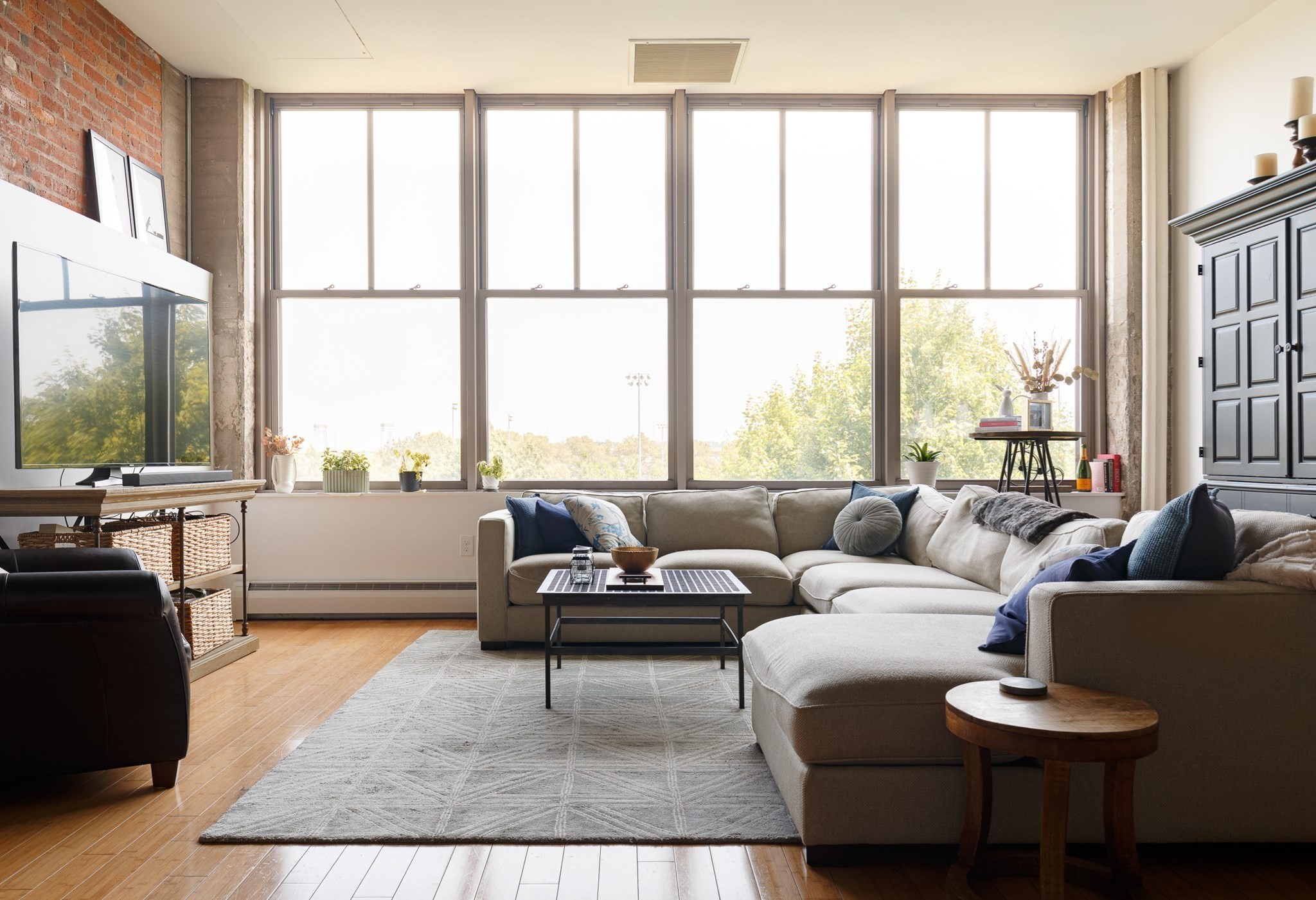Living room with a large sectional sofa, gray rug, and tall windows overlooking trees.