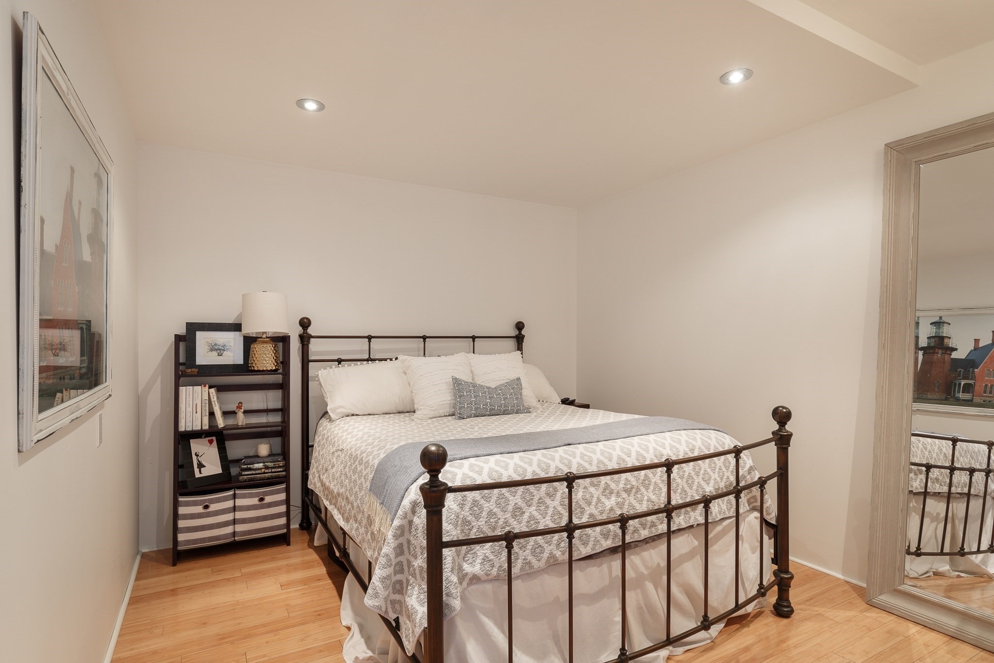 Bedroom with a dark metal bed, pale patterned bedding, hardwood floors, and a full-length mirror.