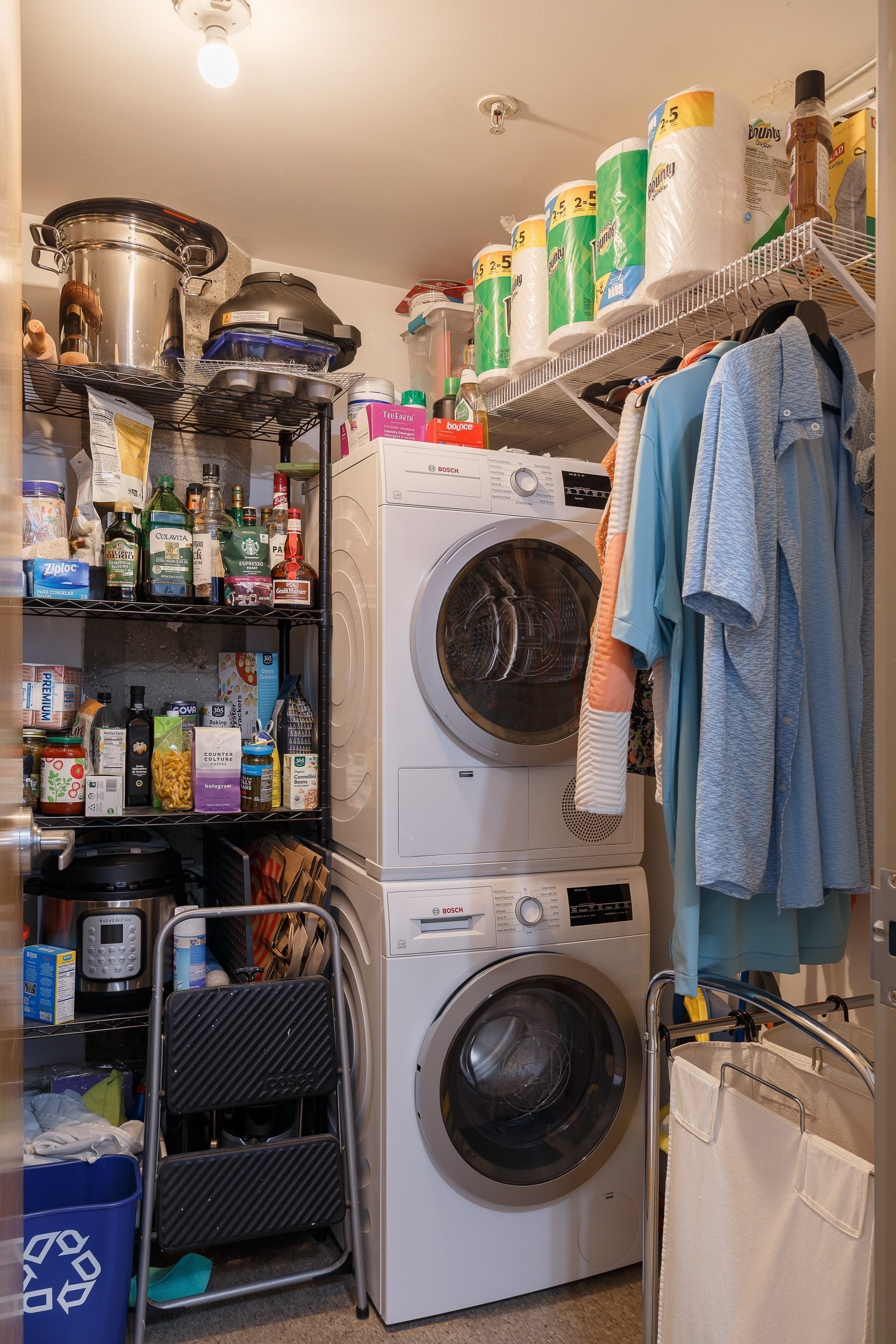 Laundry and storage room with a stacked washer and dryer, crowded shelves, and clothes hanging nearby.