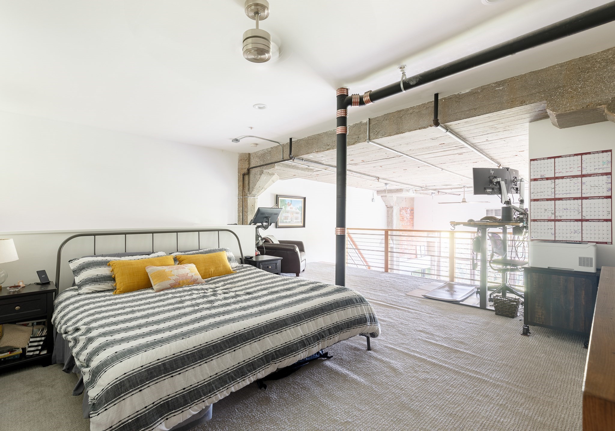 Loft bedroom with a striped bedspread, yellow pillows, exposed beams and pipes, and a workspace beyond the railing.