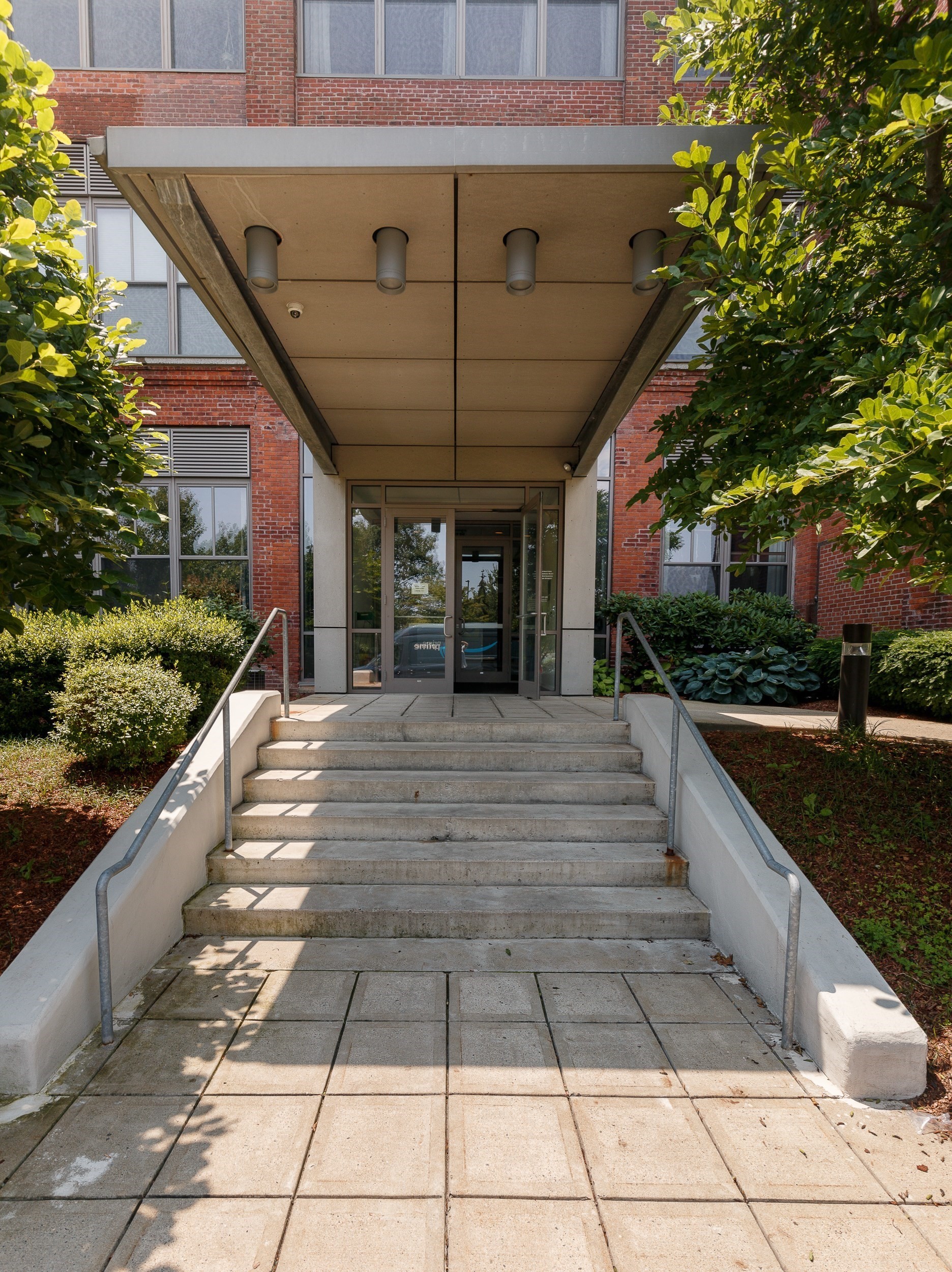 Apartment building entrance with brick facade, broad canopy, concrete steps, and leafy landscaping.