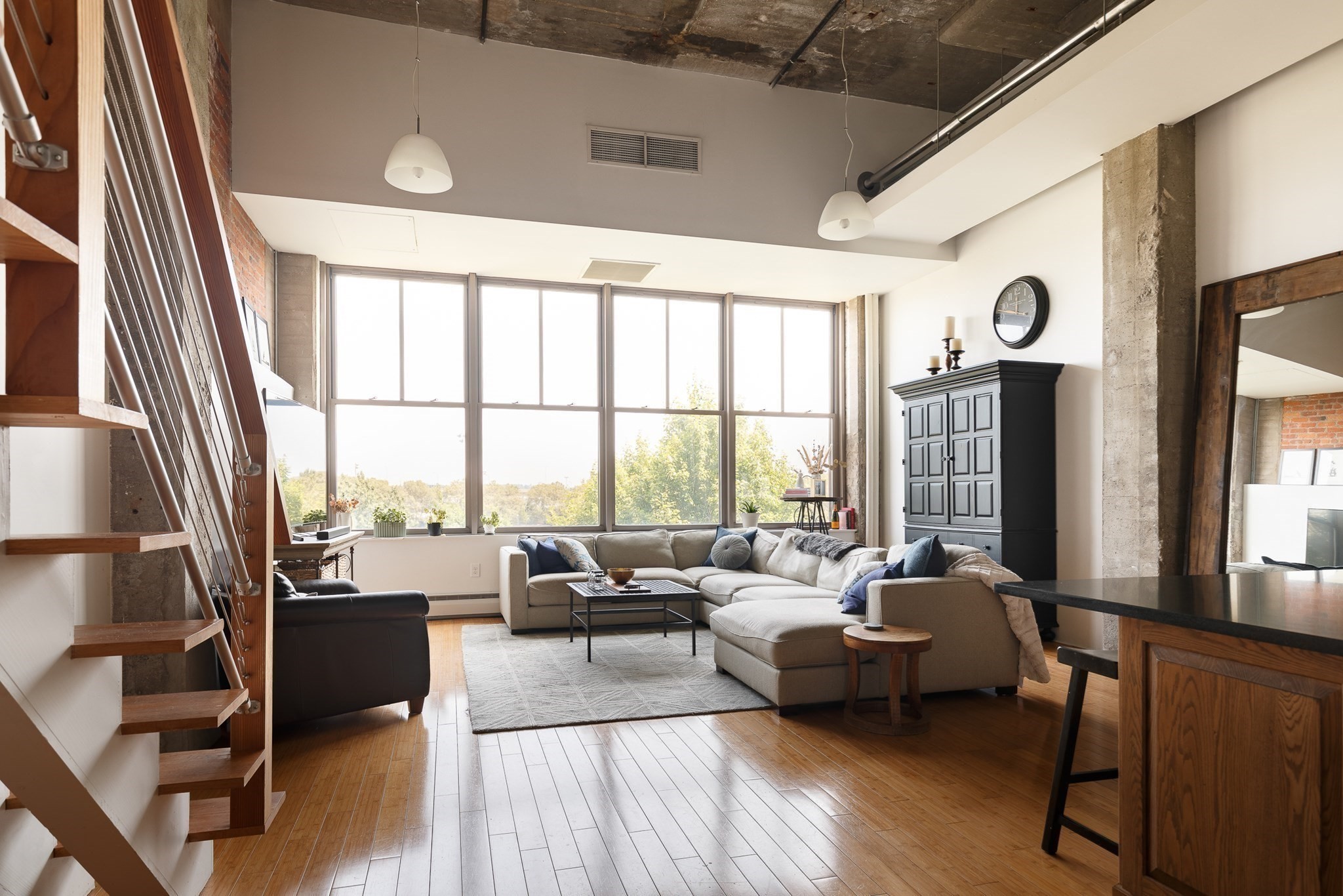 Loft living room with a gray sectional, tall windows, exposed concrete ceiling, and open-riser staircase.