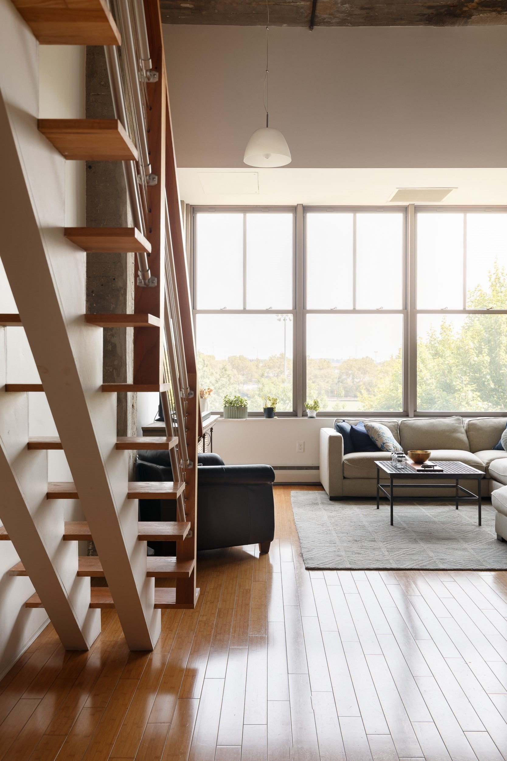 Living room with an open-riser staircase, gray sofa, and tall windows overlooking trees.