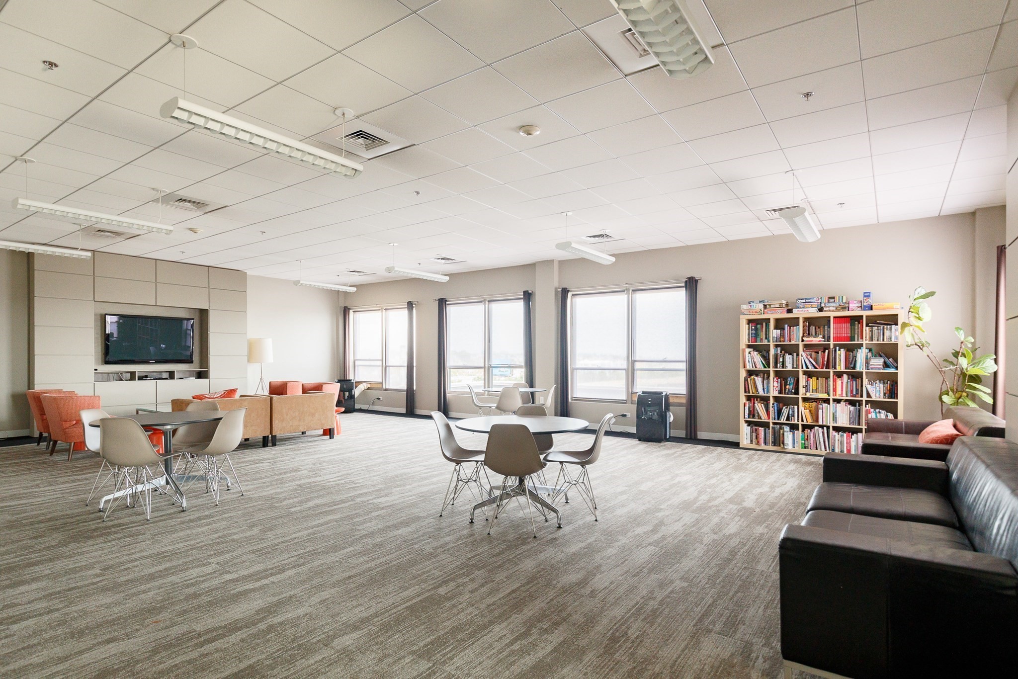 Furnished communal lounge with seating, dining tables, a bookcase, and large windows.