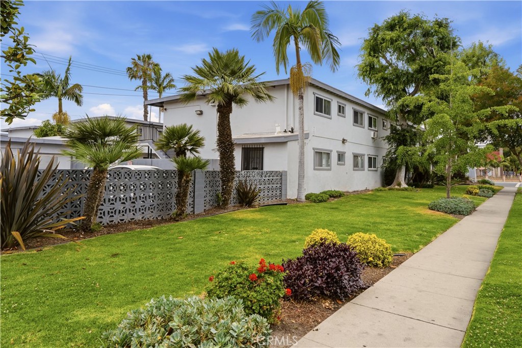 Two-story white residential building behind palm trees, a patterned fence, and a landscaped lawn.
