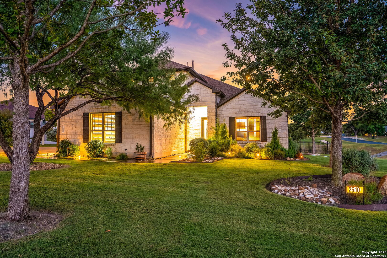 Stone house at dusk with warm window lights, mature trees, and an illuminated 2610 address marker.