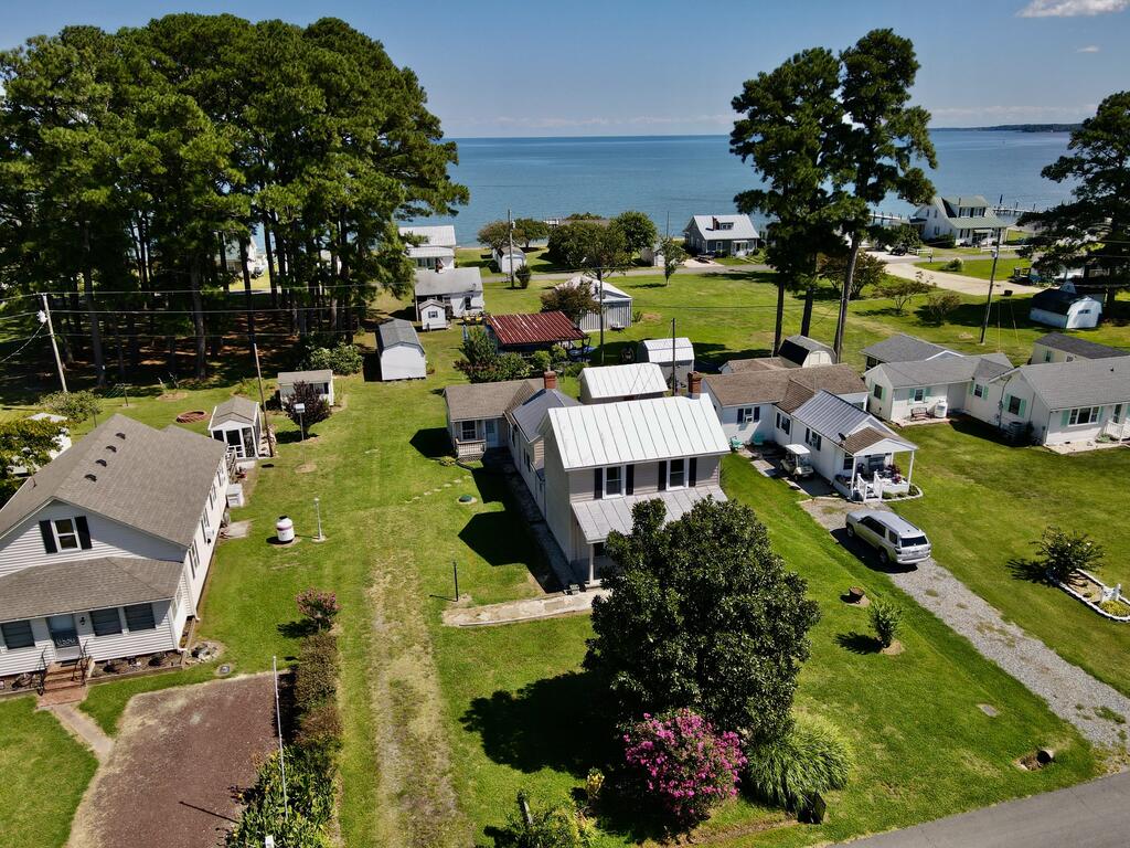 Aerial view of a light-roofed home among waterfront cottages, with mature trees and water beyond.