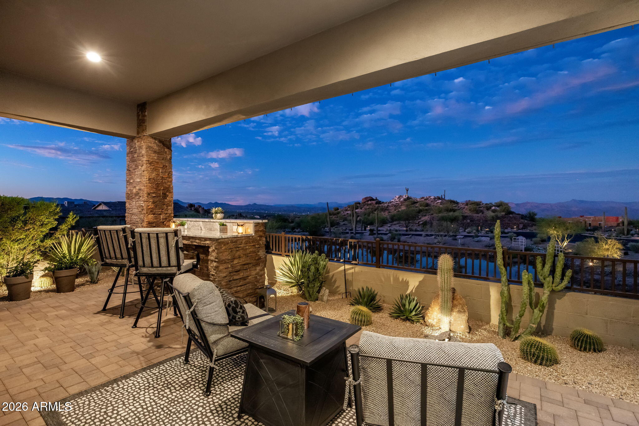 Covered patio with stone fireplace, desert landscaping, pool, and distant mountain views at dusk.