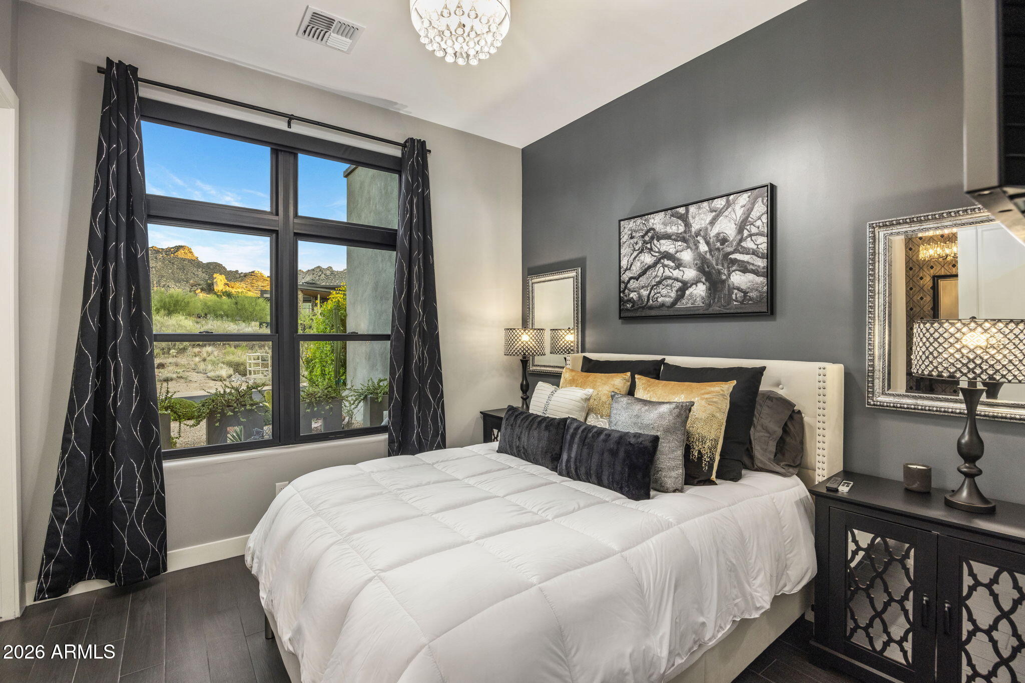 Furnished bedroom with a white bed and large window overlooking desert vegetation and rocky mountains.