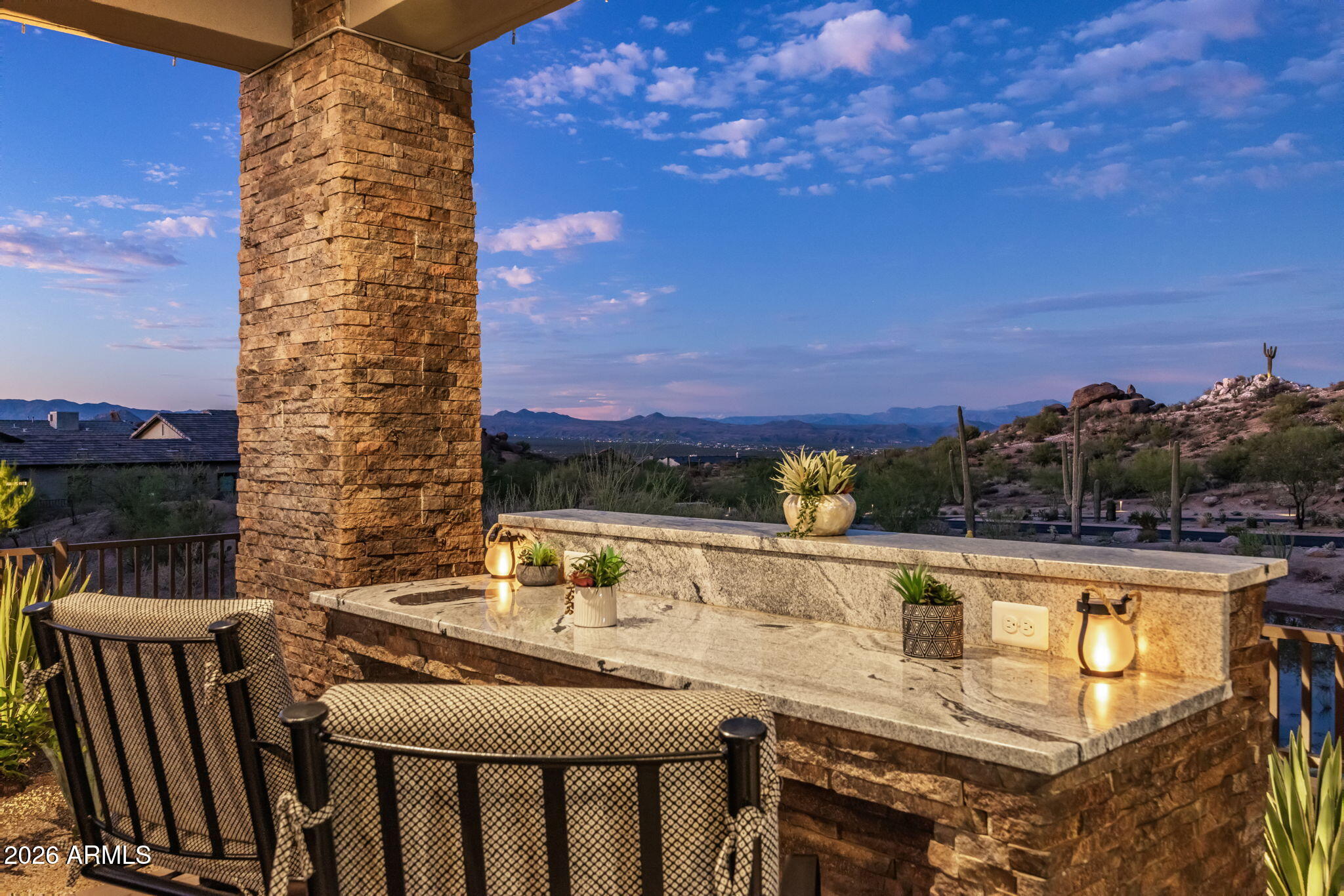 Covered stone patio with cushioned seating and lanterns overlooking desert hills and distant mountains.