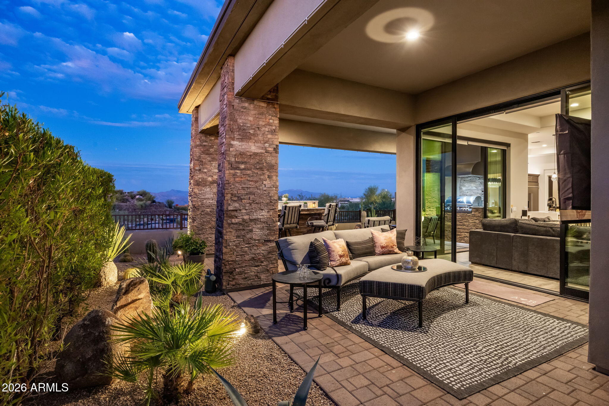 Covered patio with cushioned seating, desert landscaping, and distant mountain views at dusk.