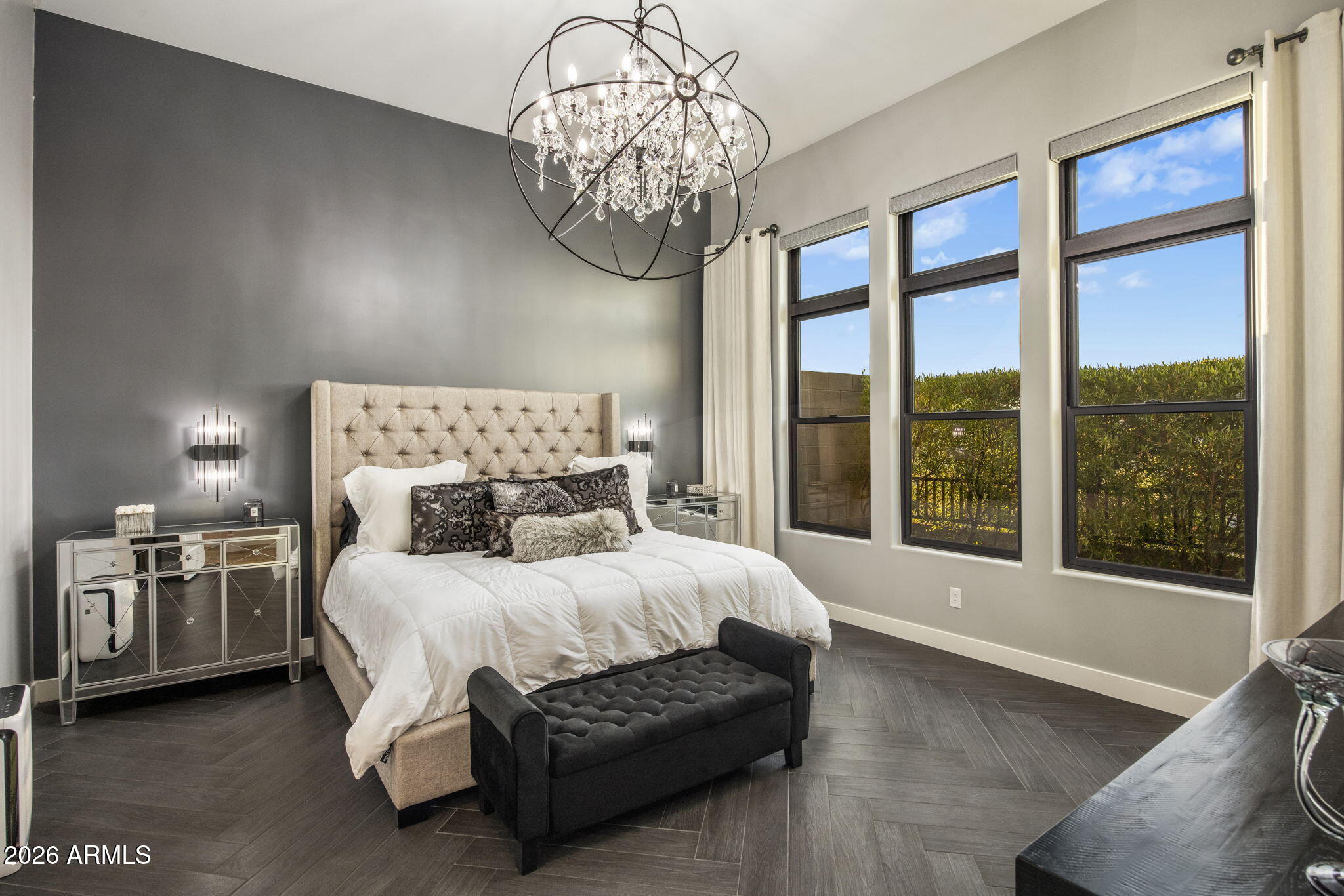 Bedroom with a tufted beige headboard, white bedding, crystal chandelier, and tall windows.