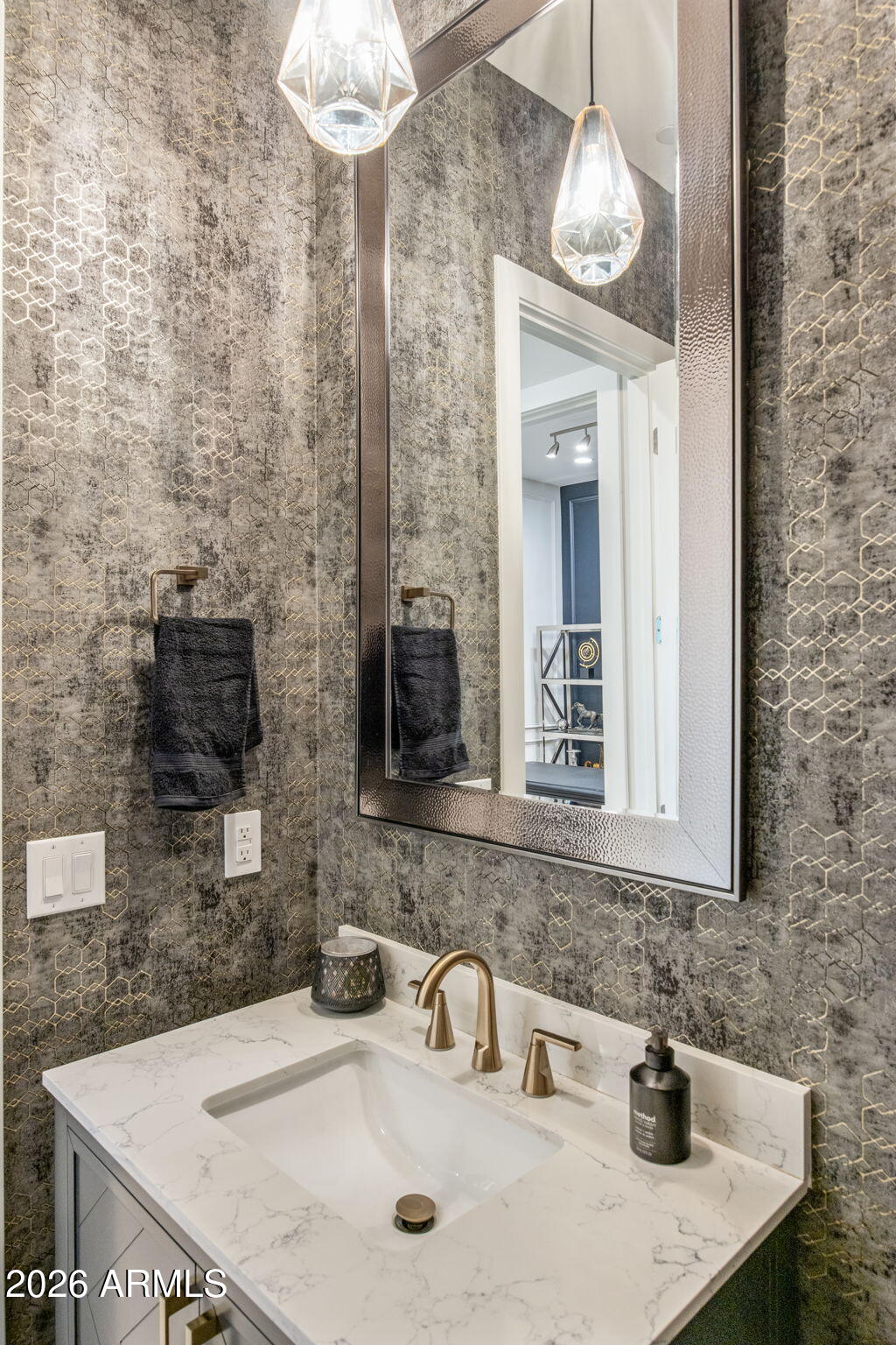 Bathroom vanity with brass faucet, marble-look counter, framed mirror, and geometric wallpaper.