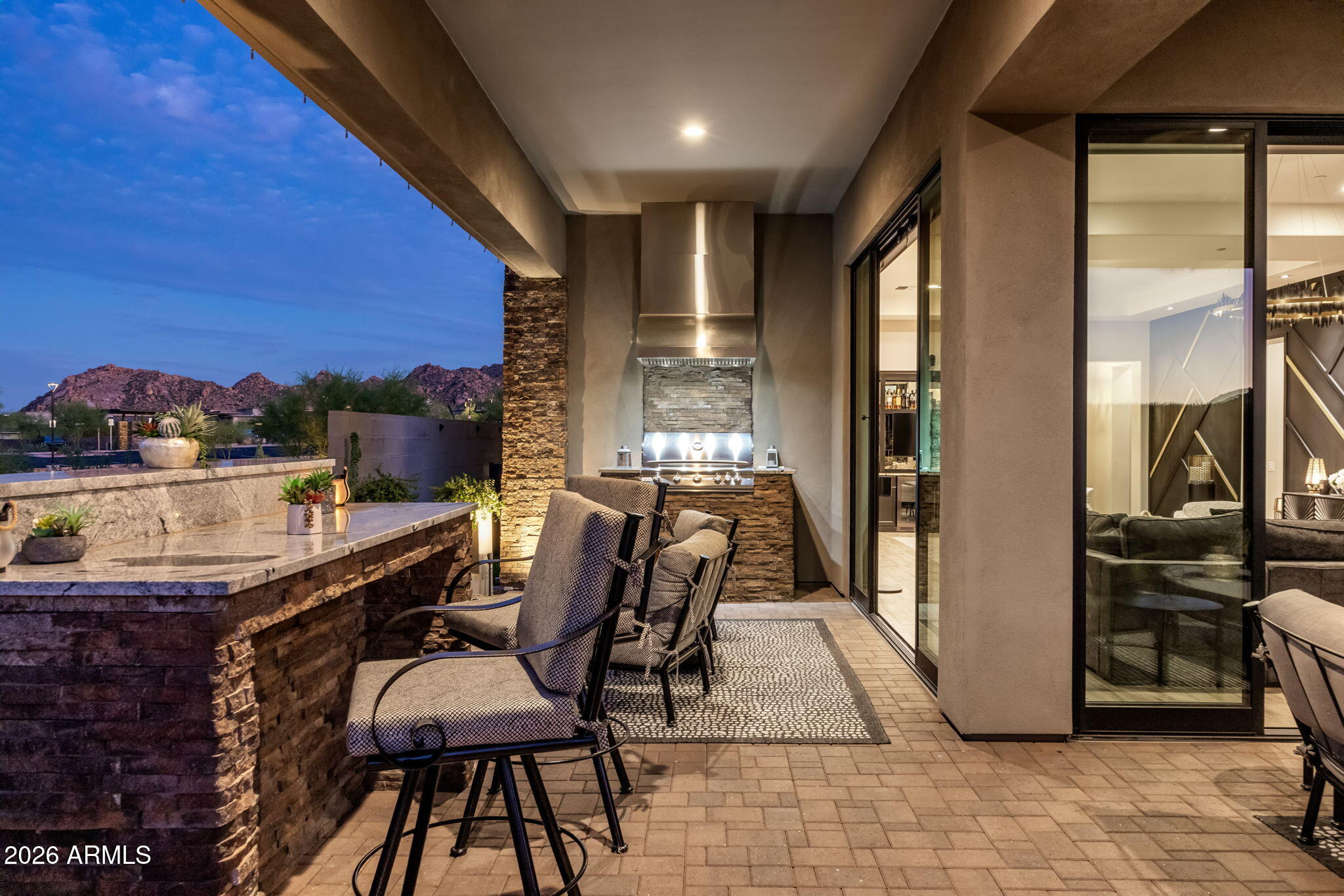 Covered patio with built-in grill, stone bar counter, cushioned seating, and mountain views at dusk.