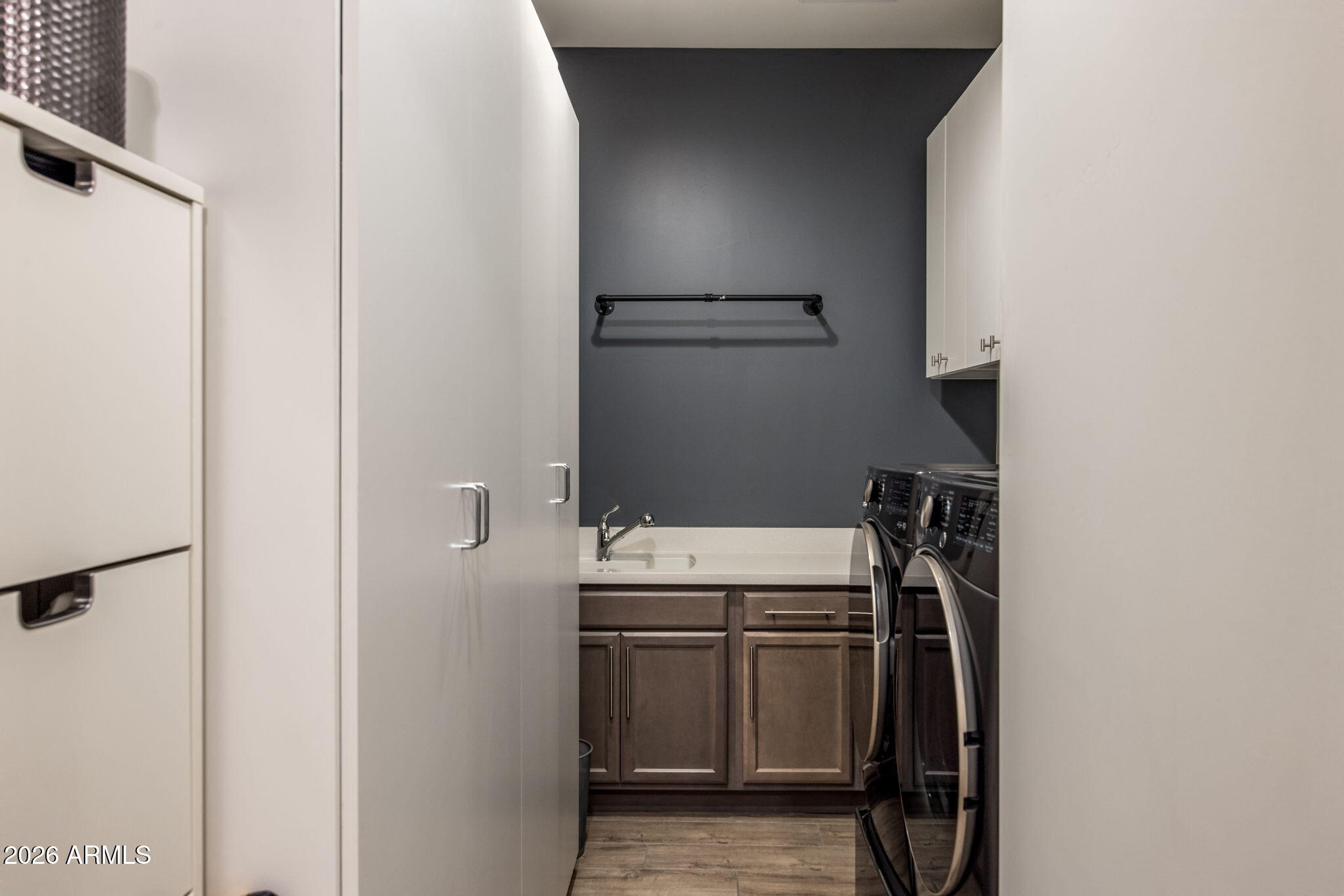 Laundry room with front-loading washer and dryer, utility sink, and white and dark wood cabinets.