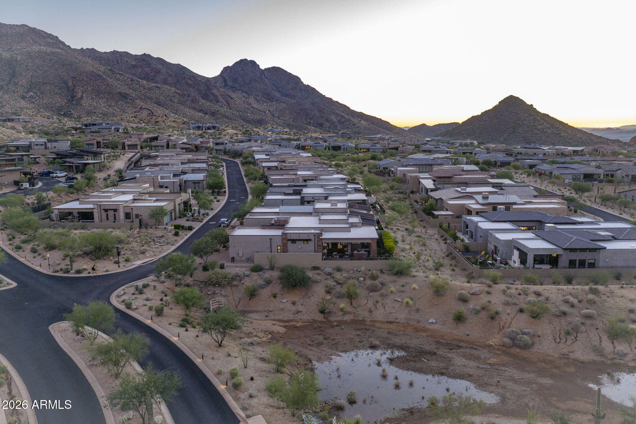 Aerial view of desert homes beneath rugged mountains at dusk, with curving neighborhood streets.